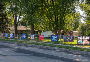 A row of political signs displayed along a residential fence during the 2024 U.S. election cycle. The banners, supporting former President Donald Trump and expressing opposition to President Joe Biden, reflect the increasing visibility of partisan messaging in everyday public and suburban spaces. Throughout the Upper Midwest and many parts of the country, large-format yard signs and roadside banners became common ways for homeowners to express political allegiance ahead of the election.

The use of oversized, commercially printed campaign signs grew significantly after the 2016 and 2020 elections, coinciding with the rise of direct-to-consumer political merchandise and online storefronts selling non-official campaign materials. Residential displays like this illustrate how political identity has increasingly blended into street-level visual culture, especially in communities where private property is used as a platform for ongoing political messaging outside traditional election windows.