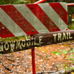 An aging “Snowmobile Trail” sign stands along a forested route in Stony River Township in northern Minnesota, an area threaded with seasonal trails maintained for winter travel. Much of this region lies within or near the Superior National Forest, where snowmobile corridors connect remote townships, logging roads, and unpaved access routes used throughout the winter months. Signs like this—often weathered, lichen-covered, and mounted on repurposed road barricades—reflect the long history of recreational and utility snowmobiling in northern Minnesota, where deep snow and long winters have shaped a network of informal and designated trails since the mid-20th century.