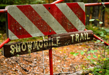 An aging “Snowmobile Trail” sign stands along a forested route in Stony River Township in northern Minnesota, an area threaded with seasonal trails maintained for winter travel. Much of this region lies within or near the Superior National Forest, where snowmobile corridors connect remote townships, logging roads, and unpaved access routes used throughout the winter months. Signs like this—often weathered, lichen-covered, and mounted on repurposed road barricades—reflect the long history of recreational and utility snowmobiling in northern Minnesota, where deep snow and long winters have shaped a network of informal and designated trails since the mid-20th century.