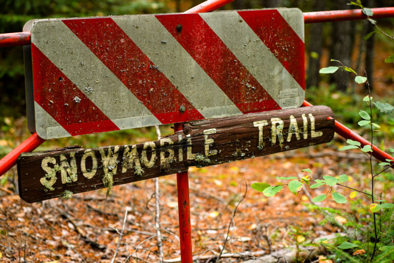 Snowmobile Trail Sign in Stony River Township 2 An aging “Snowmobile Trail” sign stands along a forested route in Stony River Township in northern Minnesota, an area threaded with seasonal trails maintained for winter travel. Much of this region lies within or near the Superior National Forest, where snowmobile corridors connect remote townships, logging roads, and unpaved access routes used throughout the winter months. Signs like this—often weathered, lichen-covered, and mounted on repurposed road barricades—reflect the long history of recreational and utility snowmobiling in northern Minnesota, where deep snow and long winters have shaped a network of informal and designated trails since the mid-20th century.