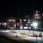A nighttime view of the USG industrial plant glows under rows of bright sodium and LED security lights. The red-sided facility’s sign stands out sharply in white and red against the dark sky, while the complex’s metallic towers and piping shimmer under spotlights in the background. The quiet lot, railway siding, and small illuminated guard booth in the foreground add a sense of stillness to an otherwise powerful industrial scene — a snapshot of modern manufacturing infrastructure at rest, yet poised for constant operation.