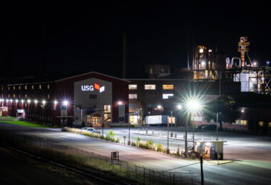A nighttime view of the USG industrial plant glows under rows of bright sodium and LED security lights. The red-sided facility’s sign stands out sharply in white and red against the dark sky, while the complex’s metallic towers and piping shimmer under spotlights in the background. The quiet lot, railway siding, and small illuminated guard booth in the foreground add a sense of stillness to an otherwise powerful industrial scene — a snapshot of modern manufacturing infrastructure at rest, yet poised for constant operation.
