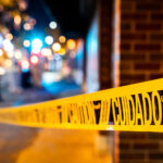 A length of bright yellow cuidado (caution) tape stretches across a sidewalk in Minneapolis, marking off an area temporarily closed to pedestrians. These barriers are a common sight during routine municipal work, building maintenance, or street-level repairs, especially in dense commercial districts where crews often work overnight. Blurred streetlights and storefront colors in the background reflect the activity of the surrounding neighborhood and the way safety markers become part of the visual landscape of city life.