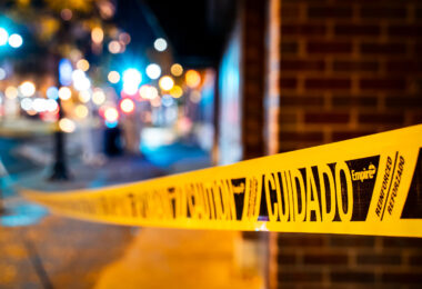 A length of bright yellow cuidado (caution) tape stretches across a sidewalk in Minneapolis, marking off an area temporarily closed to pedestrians. These barriers are a common sight during routine municipal work, building maintenance, or street-level repairs, especially in dense commercial districts where crews often work overnight. Blurred streetlights and storefront colors in the background reflect the activity of the surrounding neighborhood and the way safety markers become part of the visual landscape of city life.