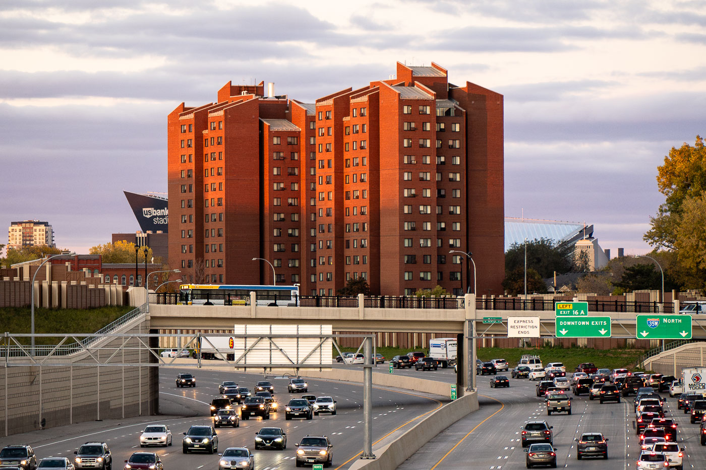 The Fifth Avenue High-Rise, a Minneapolis Public Housing Authority building completed in the early 1970s, stands just east of downtown and overlooks the busy stretch of Interstate 35W seen below. Designed during an era when high-density towers were a common approach to urban public housing, the structure is part of a cluster of residential buildings intended to provide affordable units for seniors and residents with disabilities. In this view from October 2021, the west-facing façade catches the warm late-day light while evening traffic builds on I-35W toward downtown. In the background, the angular roofline of U.S. Bank Stadium marks the eastern edge of the central business district, situating the high-rise within one of the city’s most infrastructure-dense corridors.