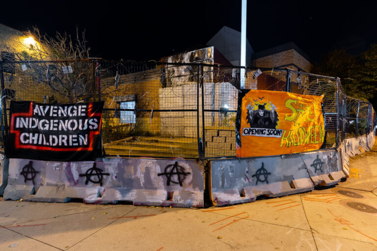 Minneapolis Police 3rd Precinct: Spirit Halloween Banners on Burned Building 2 Banners reading "Avenge Indigenous Children" and "Spirit Halloween" are displayed on the barricaded perimeter of the former Minneapolis Police Third Precinct. The building was set ablaze during protests that erupted following the murder of George Floyd on May 25, 2020. The precinct, located in the Powderhorn Park neighborhood, was a focal point of unrest and has remained a symbol of the protests against police brutality. The "Spirit Halloween" banner, featuring a grim reaper figure and the text "Opening Soon!", was placed on the site after the building's destruction.