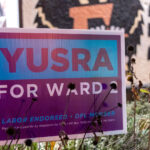 A campaign sign for Yusra Arab stands among late-season plants in Minneapolis’ Ward 2, one of the city’s most politically active and demographically diverse districts. Ward 2 includes neighborhoods such as Cedar-Riverside, Prospect Park, Seward, and the University district—areas shaped by long histories of immigration, student populations, and tenant-driven community organizing. Yard signs like this became common during the municipal election cycle as candidates focused on issues that have defined Ward 2 politics for decades: rental housing policy, transportation infrastructure, campus-area safety, and the evolving relationship between the University of Minnesota and surrounding neighborhoods.

Minneapolis’ ward system has historically produced competitive races, especially in districts with high levels of civic engagement and shifting demographics. Ward 2 is known for its strong DFL presence, multi-lingual community outreach, and a mix of long-term residents and new arrivals. Campaign messaging in the ward often reflects its unique blend of grassroots activism, student-led political energy, and deep neighborhood-level involvement in city governance.