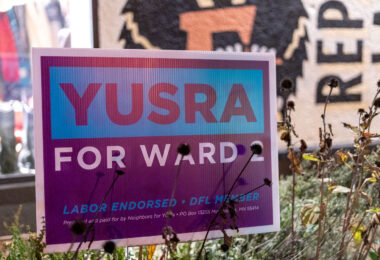 A campaign sign for Yusra Arab stands among late-season plants in Minneapolis’ Ward 2, one of the city’s most politically active and demographically diverse districts. Ward 2 includes neighborhoods such as Cedar-Riverside, Prospect Park, Seward, and the University district—areas shaped by long histories of immigration, student populations, and tenant-driven community organizing. Yard signs like this became common during the municipal election cycle as candidates focused on issues that have defined Ward 2 politics for decades: rental housing policy, transportation infrastructure, campus-area safety, and the evolving relationship between the University of Minnesota and surrounding neighborhoods.

Minneapolis’ ward system has historically produced competitive races, especially in districts with high levels of civic engagement and shifting demographics. Ward 2 is known for its strong DFL presence, multi-lingual community outreach, and a mix of long-term residents and new arrivals. Campaign messaging in the ward often reflects its unique blend of grassroots activism, student-led political energy, and deep neighborhood-level involvement in city governance.