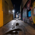 This narrow Uptown alleyway comes alive after dark with the vibrant work of local artist Black Daze, whose murals brighten the service corridors behind Hennepin Avenue’s bars, restaurants, and small storefronts. The deep colors, stylized florals, and swirling patterns contrast sharply with the utilitarian yellow brick wall opposite, highlighting the way Minneapolis’ alleys often double as informal public art galleries. Puddles from a recent rain reflect the overhead security lights, adding texture to a space normally overlooked in the bustle of Uptown’s nightlife.