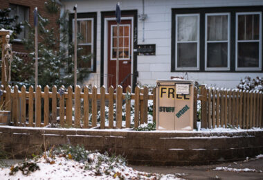 A small handmade “Free Stuff Inside” cabinet sits beside a picket fence in a residential Minneapolis neighborhood, offering passersby clothing, household items, and other essentials at no cost. These volunteer-maintained boxes—modeled loosely after Little Free Pantries and similar mutual-aid projects—became common across the city in recent years as neighbors looked for practical ways to support one another. This particular cabinet shows signs of winter use, with a thin layer of snow on the ground and along the fence, emphasizing the year-round effort by residents to keep shared resources accessible.

The rise of these informal exchange boxes reflects a broader culture of grassroots support systems that grew during the pandemic and the 2020 unrest. Neighbors created hyper-local networks for food, winter gear, and daily necessities, often improvising with whatever materials they had. Even a small cabinet like this served as a reliable touchpoint for people needing gloves, toiletries, or household supplies, helping strengthen the sense of community resilience in the city’s residential blocks.