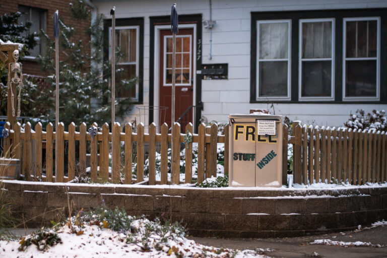 Neighborhood Free-Stuff Cabinet in Winter 1 A small handmade “Free Stuff Inside” cabinet sits beside a picket fence in a residential Minneapolis neighborhood, offering passersby clothing, household items, and other essentials at no cost. These volunteer-maintained boxes—modeled loosely after Little Free Pantries and similar mutual-aid projects—became common across the city in recent years as neighbors looked for practical ways to support one another. This particular cabinet shows signs of winter use, with a thin layer of snow on the ground and along the fence, emphasizing the year-round effort by residents to keep shared resources accessible.The rise of these informal exchange boxes reflects a broader culture of grassroots support systems that grew during the pandemic and the 2020 unrest. Neighbors created hyper-local networks for food, winter gear, and daily necessities, often improvising with whatever materials they had. Even a small cabinet like this served as a reliable touchpoint for people needing gloves, toiletries, or household supplies, helping strengthen the sense of community resilience in the city’s residential blocks.