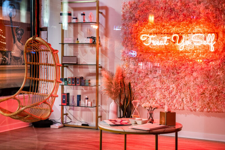 Treat Yo Self Neon Sign, Lake Street, Minneapolis 1 A neon sign reading "Treat Yo Self" illuminates a retail space on Lake Street in Minneapolis. The sign is mounted on a wall covered in artificial pink flowers, creating a vibrant and inviting atmosphere for customers. The surrounding decor, including a hanging rattan chair and shelves stocked with beauty products, suggests the establishment is a boutique or spa. Lake Street is a major commercial corridor in Minneapolis, known for its diverse businesses and cultural significance.
