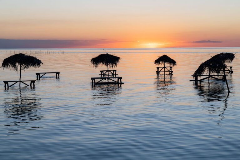 Beautiful sunset at Secret Beach Ambergris Caye, Belize 2 A calm evening sunset over the shallow waters of Secret Beach on Ambergris Caye, Belize. The wooden overwater tables and palapa-style shade canopies sit just offshore, a distinctive feature of this west-facing stretch of the island where the coastline opens onto the protected waters of Ambergris Bay. As the sun drops toward the horizon, the sky reflects soft orange and pink tones across the glassy surface of the bay, highlighting the area’s reputation as one of the island’s most popular sunset viewpoints.