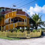 Casa Johnson on Ambergris Caye 1 Casa Johnson stands along one of the sandy backroads north of San Pedro, a brightly painted elevated home typical of the island’s hurricane-resilient architecture. The structure’s sweeping exterior staircase and open upper-level deck reflect a style common to private residences and small guest properties built for cross-breezes and views toward the Caribbean Sea. Homes in this area of Ambergris Caye often sit on raised foundations to protect against storm surge and seasonal flooding, a practical requirement on the low-lying island. The neighborhood around Casa Johnson has grown steadily over the past two decades, transitioning from scattered beachfront lots into a mix of long-term residences, rental units, and small locally owned businesses that line the northern approach to San Pedro.