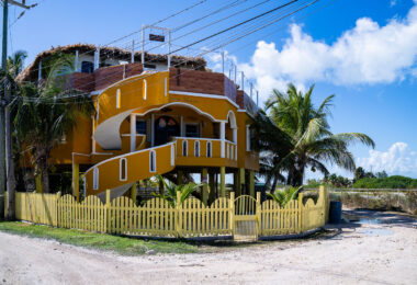 Casa Johnson stands along one of the sandy backroads north of San Pedro, a brightly painted elevated home typical of the island’s hurricane-resilient architecture. The structure’s sweeping exterior staircase and open upper-level deck reflect a style common to private residences and small guest properties built for cross-breezes and views toward the Caribbean Sea. Homes in this area of Ambergris Caye often sit on raised foundations to protect against storm surge and seasonal flooding, a practical requirement on the low-lying island. The neighborhood around Casa Johnson has grown steadily over the past two decades, transitioning from scattered beachfront lots into a mix of long-term residences, rental units, and small locally owned businesses that line the northern approach to San Pedro.