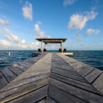 Dock Near Matachica Resort, Ambergris Caye 4 A long wooden dock stretches into the Caribbean Sea just north of Matachica Resort & Spa on Ambergris Caye, constructed in a distinctive chevron pattern common to many of the island’s piers. These docks serve as essential access points for small boats, snorkeling excursions, and water taxis, reflecting the island’s long reliance on marine travel. At the end of the pier, a shaded palapa provides a quiet spot overlooking the protected waters inside the Belize Barrier Reef, where shallow seagrass flats and calm tides have supported fishing communities and tourism infrastructure for decades.