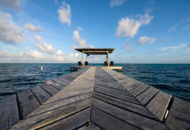 A long wooden dock stretches into the Caribbean Sea just north of Matachica Resort & Spa on Ambergris Caye, constructed in a distinctive chevron pattern common to many of the island’s piers. These docks serve as essential access points for small boats, snorkeling excursions, and water taxis, reflecting the island’s long reliance on marine travel. At the end of the pier, a shaded palapa provides a quiet spot overlooking the protected waters inside the Belize Barrier Reef, where shallow seagrass flats and calm tides have supported fishing communities and tourism infrastructure for decades.