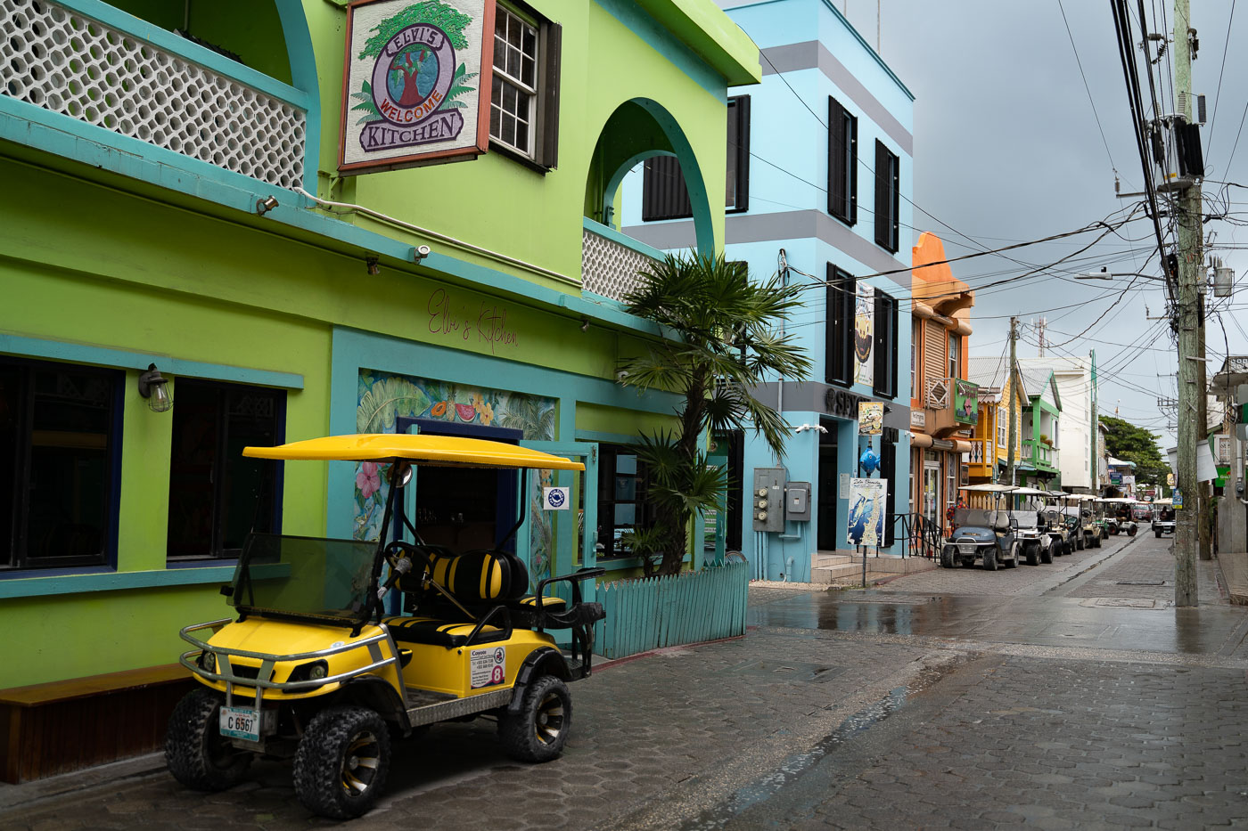 Golf carts line a cobblestone street in San Pedro, Belize, in front of Elvi's Kitchen restaurant under an overcast sky.