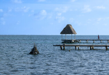 A wooden pier extends into the Caribbean Sea on the eastern shore of Ambergris Caye, topped with a palapa-style shelter commonly found along the island’s waterfront. These simple wooden structures serve both locals and visitors, offering space for fishing, small-boat landings, and relief from the midday sun. Just offshore, the shallow coastal waters give way to the darker blue of the open sea, with the white line of breakers marking the crest of the Belize Barrier Reef—the second-largest reef system in the world and a central part of the island’s history, economy, and marine culture.