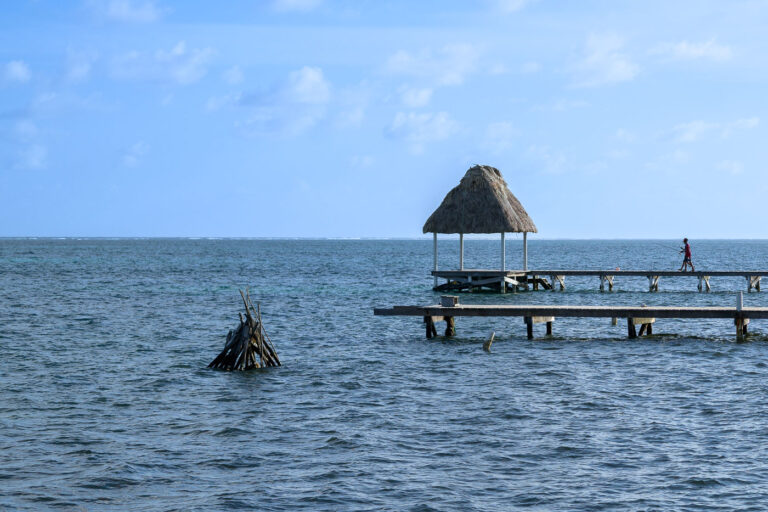 A wooden pier extends into the Caribbean Sea on the eastern shore of Ambergris Caye, topped with a palapa-style shelter commonly found along the island’s waterfront. These simple wooden structures serve both locals and visitors, offering space for fishing, small-boat landings, and relief from the midday sun. Just offshore, the shallow coastal waters give way to the darker blue of the open sea, with the white line of breakers marking the crest of the Belize Barrier Reef—the second-largest reef system in the world and a central part of the island’s history, economy, and marine culture.