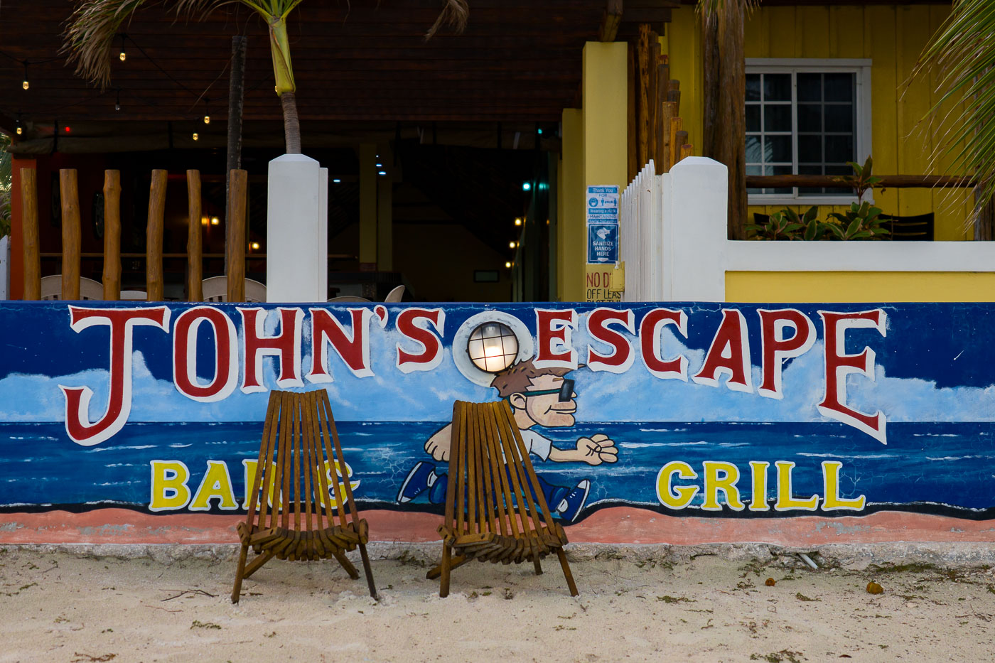 John's Escape Bar & Grill on Ambergris Caye, Belize, formerly owned by John McAfee. The establishment is situated on a beach with two wooden chairs in front of its mural.