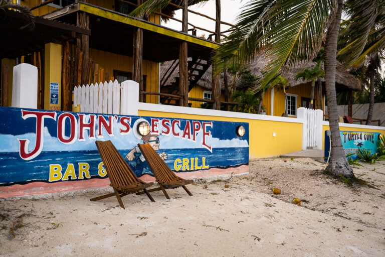 John's Escape Bar & Grill Sign 4 John’s Escape Bar & Grill sits along the sandy shoreline of Ambergris Caye, framed by palms and bright Caribbean colors that reflect the island’s casual, beach-town character. Small waterfront restaurants like this have long been part of the local coastal culture, serving residents, tour guides, and travelers arriving from nearby docks. The hand-painted mural and relaxed seating on the beach underscore how many of Ambergris Caye’s businesses blend into the natural landscape, offering views over the Belize Barrier Reef lagoon just steps from the water.