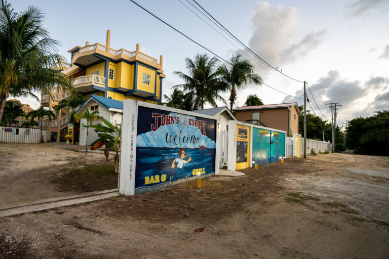 Outside John's Escape 4 A painted welcome wall marks the entrance to John’s Escape Bar & Grill on Ambergris Caye, a casual beachfront hangout located on the site once occupied by John McAfee’s former property on the island. The area has since transitioned into a small cluster of guesthouses and local businesses, but traces of its unusual past remain part of the lore in San Pedro. The restaurant sits just off the sandy road north of town, surrounded by palms, colorful buildings, and the characteristic low-rise architecture of the island’s coastal neighborhoods.