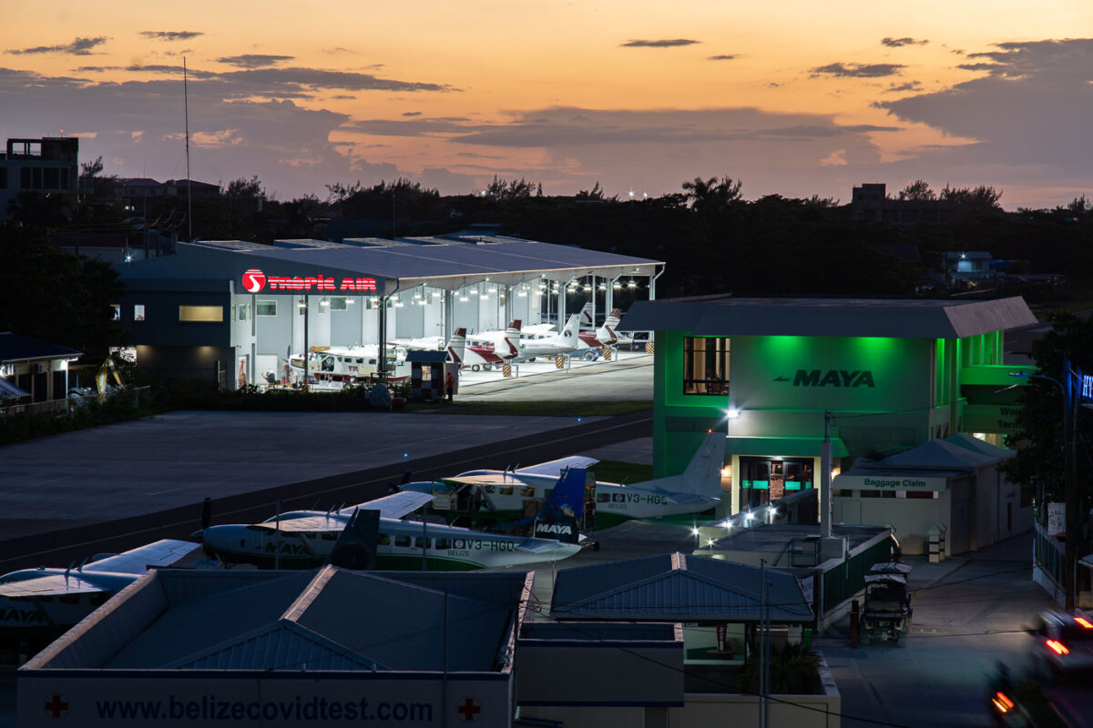 San Pedro Airport (SPR) on Ambergris Caye, Belize, at dusk, showing hangars for Tropic Air and Maya Island Air with parked aircraft.