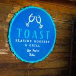 A hand-painted wooden sign marks the entrance to TOAST Seaside Boozery & Grill in San Pedro, Belize. The beachfront restaurant sits along the island’s eastern shore, an area lined with small locally owned businesses that cater to both residents and visitors exploring Ambergris Caye. The sign’s weathered texture and bright coastal colors reflect the building traditions common along the island—simple timber structures adapted to the salt air, storms, and constant humidity of the Caribbean. Establishments like TOAST contribute to the informal social landscape of San Pedro, where open-air dining rooms, beachfront swings, and walk-up bars blend directly into the surrounding beach path and waterfront access points leading toward the Belize Barrier Reef just offshore.
