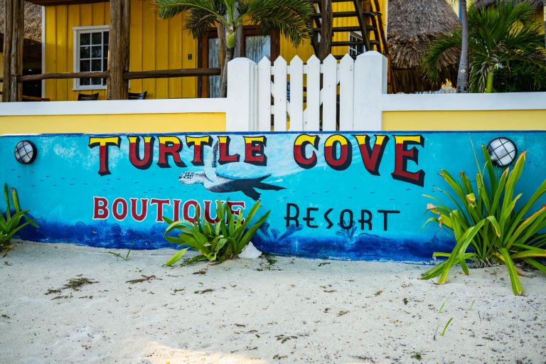Turtle Cove Boutique Resort, Ambergris Caye 1 Turtle Cove Boutique Resort sign on blue wall with yellow building and palm trees in background on Ambergris Caye, Belize.