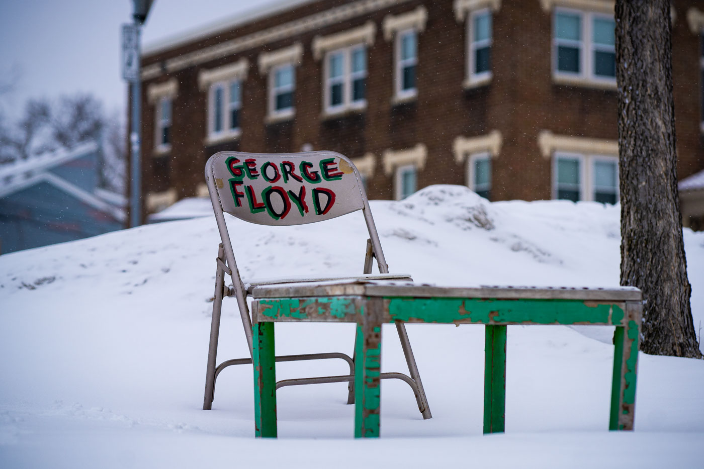A chair and table, hand-painted with "GEORGE FLOYD," are situated in the snow at George Floyd Square in Minneapolis. This location, formerly the intersection of Chicago Avenue and 38th Street, became a significant site for protests and activism following the murder of George Floyd in May 2020. The area was occupied by activists for an extended period, during which the "Say Their Names Cemetery" memorial was established. The presence of these objects in the winter landscape serves as a somber reminder of Floyd's death and the square's continued importance as a place of remembrance and protest.