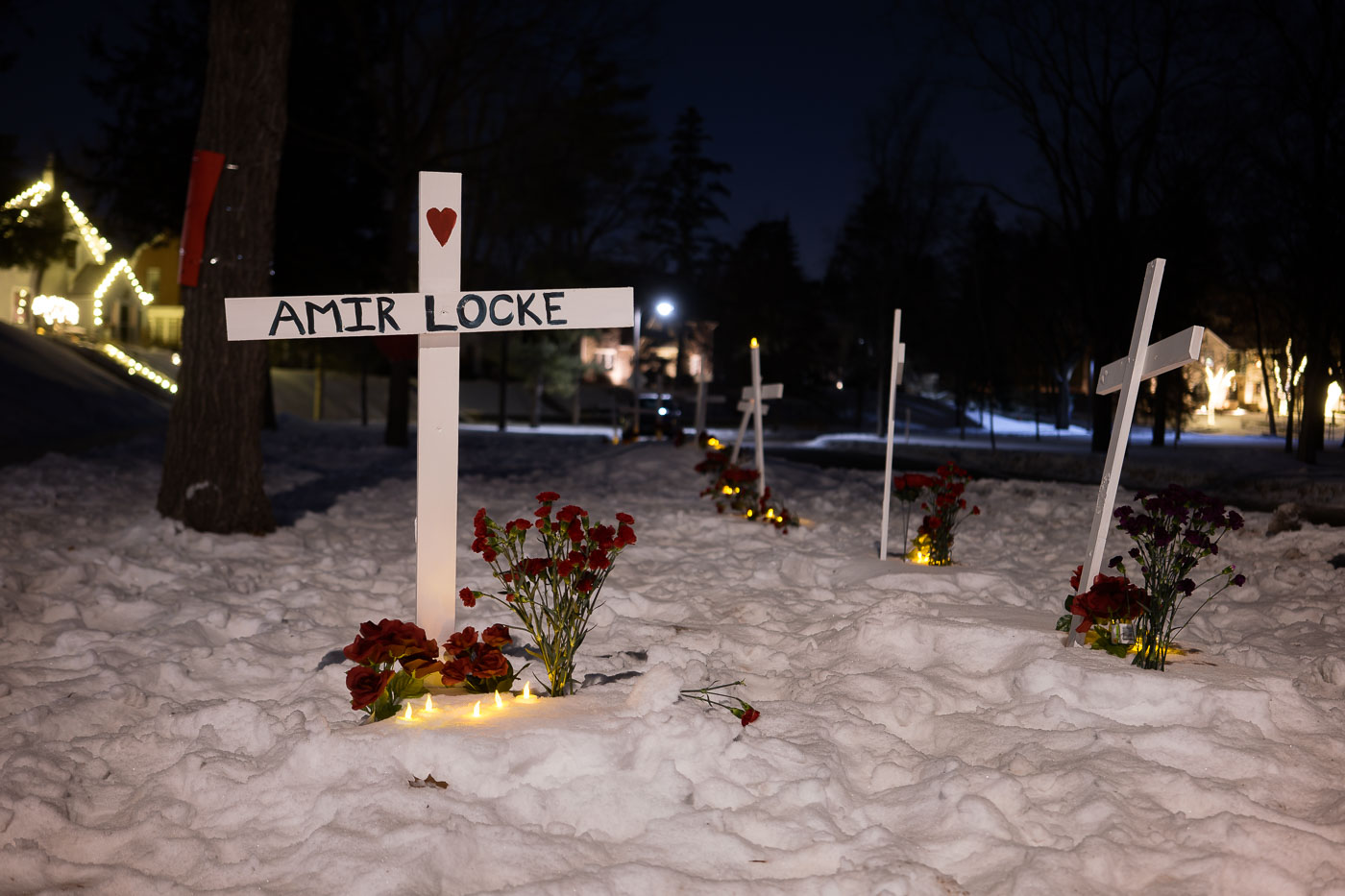 A memorial for Amir Locke, who was killed by Minneapolis police on February 2, 2022, is seen in the snow outside the home of the Minneapolis Police Chief. White crosses bearing Locke's name, along with flowers and candles, mark the site. This memorial was established following protests and community outcry over Locke's death, highlighting ongoing concerns about police accountability. The presence of multiple crosses suggests a broader memorialization of lives lost due to police violence.