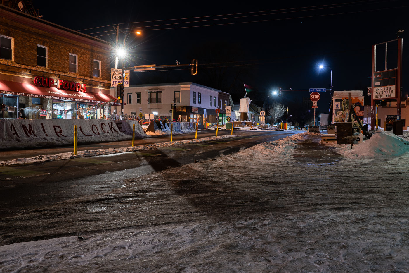 George Floyd Square in Minneapolis, a site of ongoing protest and mourning, is shown at night. Raised fist sculptures, draped in blankets in memory of Amir Locke, stand at the intersection. The area, which has been open to traffic since June, continues to serve as a community gathering space for open meetings and remembrance. The "AMIR LOCKE" banner on the barricade signifies the ongoing impact of his death on the community's activism.