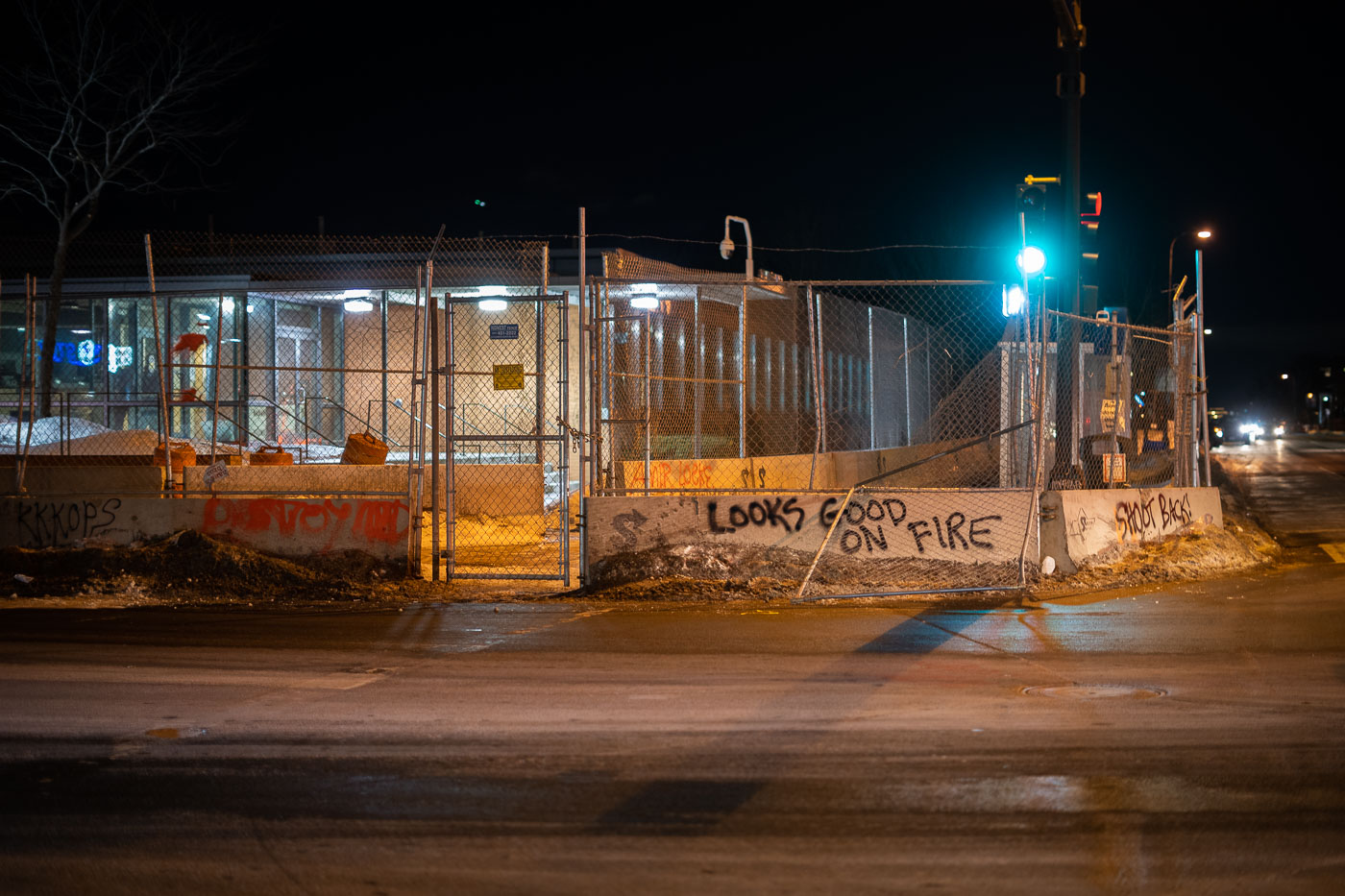 Graffiti reading "Looks Good On Fire" and "Shoot Back" is spray-painted on a construction barrier surrounding the Minneapolis Fifth Precinct building. These messages appeared during protests in February 2022, following the police shooting death of Amir Locke. The protests, which lasted for ten days, saw participants chanting "Say His Name! Amir Locke!" and leaving messages of dissent throughout the Uptown Minneapolis area. The Fifth Precinct building was under construction at the time of the events.
