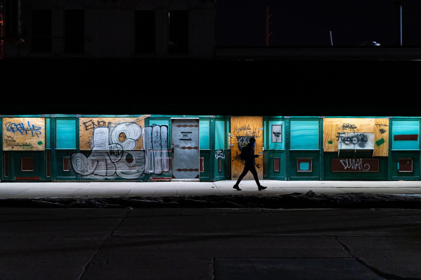 A boarded-up bar in South Minneapolis displays extensive graffiti, a visual remnant of the civil unrest that occurred in the city in 2020. The plywood covering the establishment's windows and doors is marked with various tags and messages, overlaying the building's original green facade and teal window frames. This image documents the impact of the 2020 protests on urban commercial spaces, showing a period of damage and temporary closure in a neighborhood affected by social upheaval.