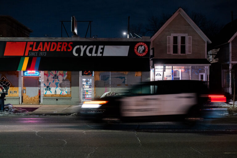 Flanders Cycle boarded up, Minneapolis Police car passes 3 Flanders Cycle, a bicycle shop established in 1973, is boarded up with plywood on Lyndale Avenue in Minneapolis. The plywood covering is a common measure taken by businesses during periods of civil unrest. A Minneapolis Police Department vehicle passes the storefront at night, reflecting the heightened tensions in the city following the murder of George Floyd in May 2020. The "BLM" graffiti visible on the plywood further signifies the context of the Minneapolis Uprising.