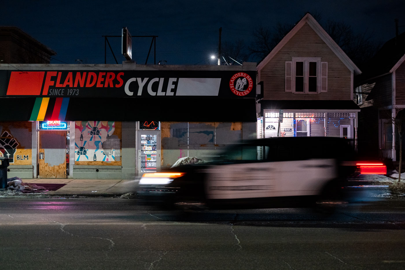 Flanders Cycle, a bicycle shop established in 1973, is boarded up with plywood on Lyndale Avenue in Minneapolis. The plywood covering is a common measure taken by businesses during periods of civil unrest. A Minneapolis Police Department vehicle passes the storefront at night, reflecting the heightened tensions in the city following the murder of George Floyd in May 2020. The "BLM" graffiti visible on the plywood further signifies the context of the Minneapolis Uprising.