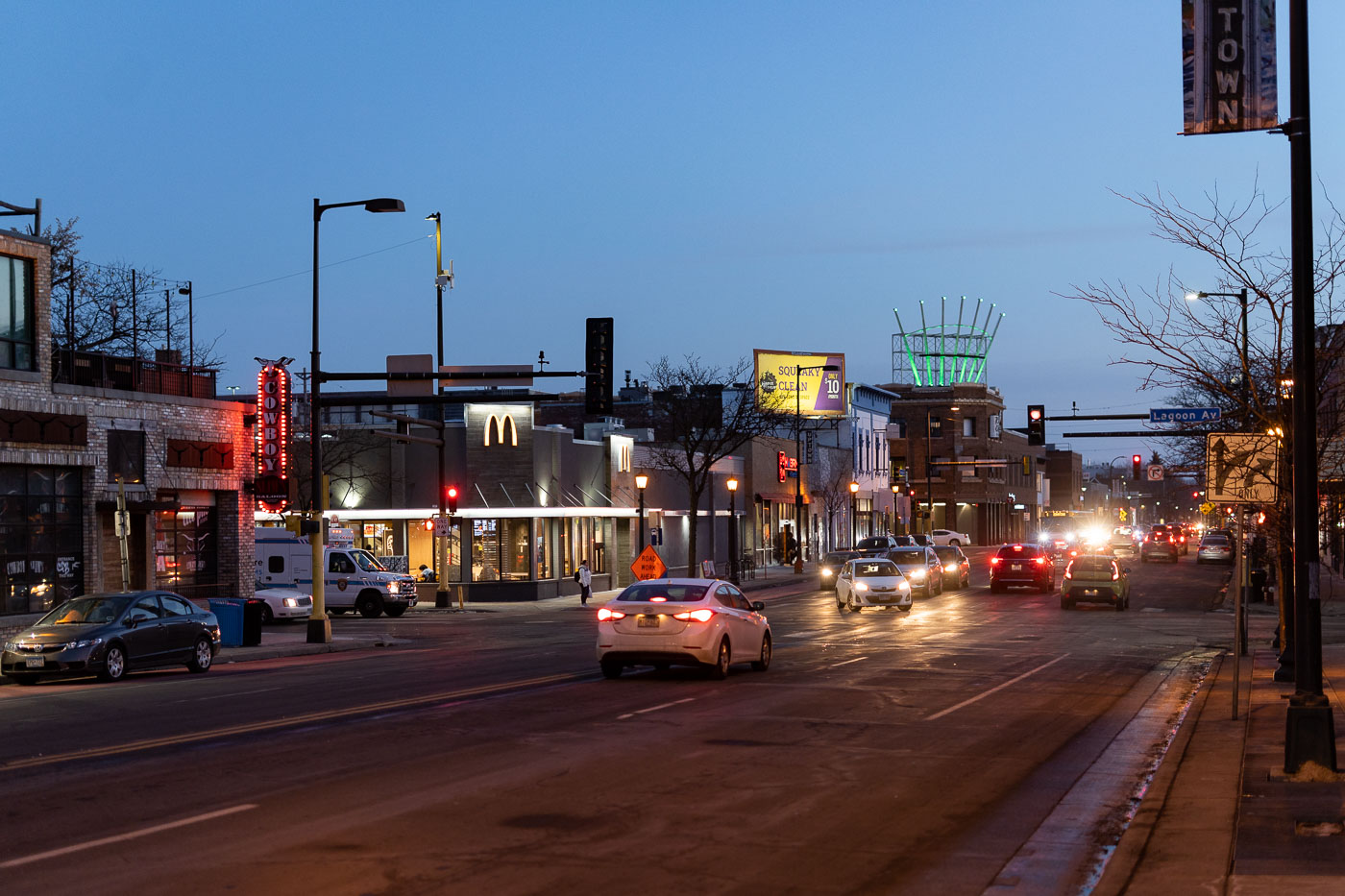 Hennepin Avenue and Lagoon Avenue in Uptown Minneapolis at dusk. Hennepin Avenue has long been a central commercial and transit artery for the Uptown neighborhood. This view shows the intersection, a hub of activity, with businesses like Cowboy Slim's and a McDonald's restaurant. The presence of streetlights and vehicle traffic highlights the area's ongoing function as a center for commerce and daily life.
