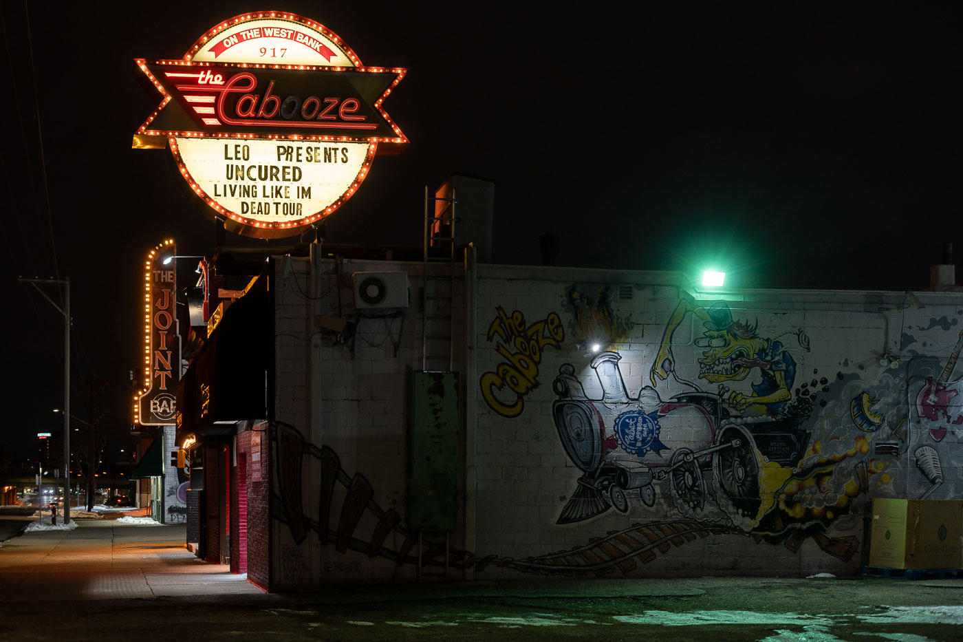 The Cabooze, a long-standing live music venue on Minneapolis's West Bank, is illuminated at night. The venue's marquee advertises a performance by Leo Presents featuring the bands Uncured and Living Like I'm Dead Tour. Established in 1969, The Cabooze has been a significant venue in the Minneapolis music scene, hosting a diverse array of local and national artists. The building's exterior features vibrant graffiti art, including a mural depicting a train engine, reflecting the venue's distinctive character and its role in the city's cultural fabric.