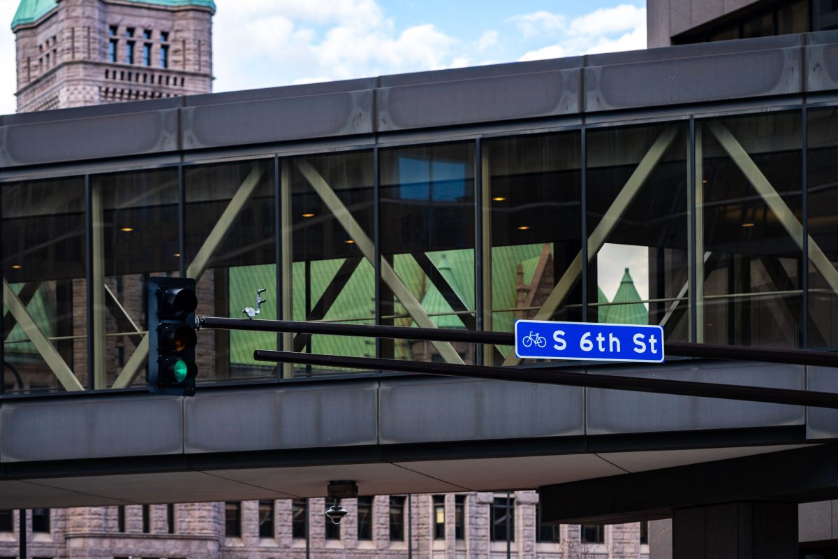 S 6th Street sign on downtown Minneapolis skyway with City Hall visible behind.