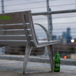 A weathered metal bench sits along one of Minneapolis’s elevated pedestrian and bicycle bridges, its frame showing rust from years of exposure to winter salt and freeze–thaw cycles. The skyline appears in the distance—soft, cool, and out of focus—giving the bench a quiet vantage point over the city. Graffiti tags on the slats and a discarded green Mickey’s bottle beneath the seat add small traces of everyday use, hinting at how these bridges serve not just as transportation corridors but as informal social spaces.

Many of the city’s long-span pedestrian bridges, especially those built over rail corridors and highways, were added during the late 20th century to connect neighborhoods divided by infrastructure. Despite their utilitarian design, they often become familiar landmarks for runners, cyclists, and residents crossing between neighborhoods at dusk, when the muted lights of downtown begin to glow against the evening sky.