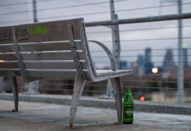 A weathered metal bench sits along one of Minneapolis’s elevated pedestrian and bicycle bridges, its frame showing rust from years of exposure to winter salt and freeze–thaw cycles. The skyline appears in the distance—soft, cool, and out of focus—giving the bench a quiet vantage point over the city. Graffiti tags on the slats and a discarded green Mickey’s bottle beneath the seat add small traces of everyday use, hinting at how these bridges serve not just as transportation corridors but as informal social spaces.

Many of the city’s long-span pedestrian bridges, especially those built over rail corridors and highways, were added during the late 20th century to connect neighborhoods divided by infrastructure. Despite their utilitarian design, they often become familiar landmarks for runners, cyclists, and residents crossing between neighborhoods at dusk, when the muted lights of downtown begin to glow against the evening sky.