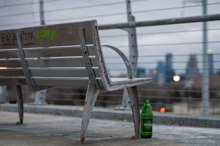 Bench Overlooking Downtown Minneapolis Mickey's Beer 2 A weathered metal bench sits along one of Minneapolis’s elevated pedestrian and bicycle bridges, its frame showing rust from years of exposure to winter salt and freeze–thaw cycles. The skyline appears in the distance—soft, cool, and out of focus—giving the bench a quiet vantage point over the city. Graffiti tags on the slats and a discarded green Mickey’s bottle beneath the seat add small traces of everyday use, hinting at how these bridges serve not just as transportation corridors but as informal social spaces.Many of the city’s long-span pedestrian bridges, especially those built over rail corridors and highways, were added during the late 20th century to connect neighborhoods divided by infrastructure. Despite their utilitarian design, they often become familiar landmarks for runners, cyclists, and residents crossing between neighborhoods at dusk, when the muted lights of downtown begin to glow against the evening sky.