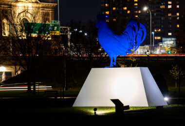 The sculpture “Hahn/Cock” by Katharina Fritsch, illuminated at night outside the Walker Art Center in Minneapolis. The ultramarine rooster, cast in fiberglass and steel, stands on a concrete plinth overlooking Hennepin Avenue and Interstate 94. Installed in 2017 as part of the Minneapolis Sculpture Garden redesign, the piece contrasts sharply against the warm façade of the Basilica of St. Mary and the light trails of evening traffic below.
