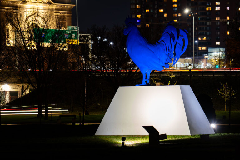 Giant Chicken sculpture by Katharina Fritsch 2 The sculpture “Hahn/Cock” by Katharina Fritsch, illuminated at night outside the Walker Art Center in Minneapolis. The ultramarine rooster, cast in fiberglass and steel, stands on a concrete plinth overlooking Hennepin Avenue and Interstate 94. Installed in 2017 as part of the Minneapolis Sculpture Garden redesign, the piece contrasts sharply against the warm façade of the Basilica of St. Mary and the light trails of evening traffic below.