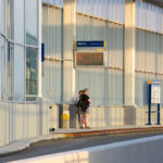 Commuters wait on the northbound platform of the I-35W & Lake Street Transit Station in Minneapolis. The elevated structure, completed in 2021 as part of the Orange Line bus rapid transit project, sits above the freeway’s center median and features glass curtain walls, enclosed waiting areas, and real-time NexTrip displays. Evening sunlight highlights the aluminum framing and sound-barrier panels that shield the station from traffic noise below.