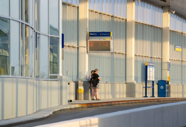Commuters wait on the northbound platform of the I-35W & Lake Street Transit Station in Minneapolis. The elevated structure, completed in 2021 as part of the Orange Line bus rapid transit project, sits above the freeway’s center median and features glass curtain walls, enclosed waiting areas, and real-time NexTrip displays. Evening sunlight highlights the aluminum framing and sound-barrier panels that shield the station from traffic noise below.