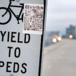A small protest sticker reading “Revolution is not a one-time event” has been placed on a “Yield to Peds” sign along a Minneapolis bike and pedestrian bridge. The bold, woodcut-style illustration echoes the city’s activist visual culture, where handmade graphics, mutual-aid posters, and political stickers have become common markers in public space since 2020. The sign stands near a long, elevated path, with downtown’s skyline softly out of focus in the distance—an everyday setting layered with the quiet reminders of ongoing organizing and civic expression.

Seen along many commuter routes, stickers like this often circulate through local art networks and community print studios, becoming part of a broader landscape of grassroots messaging that blends into the city’s infrastructure: crosswalk poles, trail markers, bridge signs, and bike lanes. The placement here reflects how Minneapolis’s trail system frequently doubles as a corridor not only for transportation but also for political communication and neighborhood identity.