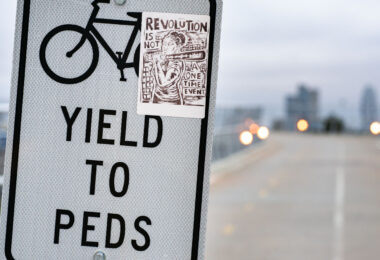 A small protest sticker reading “Revolution is not a one-time event” has been placed on a “Yield to Peds” sign along a Minneapolis bike and pedestrian bridge. The bold, woodcut-style illustration echoes the city’s activist visual culture, where handmade graphics, mutual-aid posters, and political stickers have become common markers in public space since 2020. The sign stands near a long, elevated path, with downtown’s skyline softly out of focus in the distance—an everyday setting layered with the quiet reminders of ongoing organizing and civic expression.

Seen along many commuter routes, stickers like this often circulate through local art networks and community print studios, becoming part of a broader landscape of grassroots messaging that blends into the city’s infrastructure: crosswalk poles, trail markers, bridge signs, and bike lanes. The placement here reflects how Minneapolis’s trail system frequently doubles as a corridor not only for transportation but also for political communication and neighborhood identity.