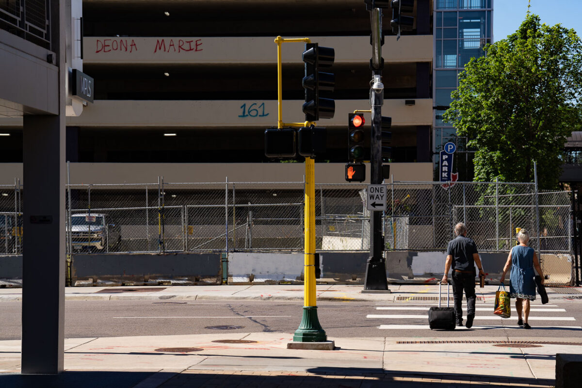 A couple walk across Lake Street in front of a parking garage with graffiti that reads “Deona Marie” “161”. The area has been a place of protest since the June 2021 killing of Winston Smith.

Smith was killed by a federal task force serving a warrant. Protester Deona Marie was killed ten days later when a man drove his vehicle through barricades killing her.